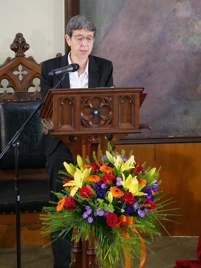 Pastor Daniela giving a sermon at the alter with a large bouquet of flowers in front of the pulpit.