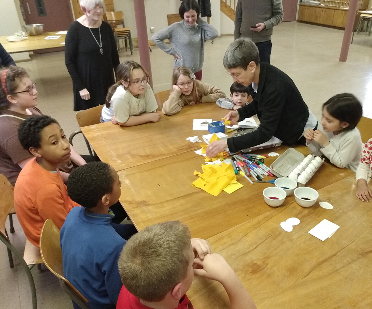 Pastor Dani is with a group of children that are sitting around a wooden table. There are eggs and dye and markers in the middle of the table for Easter egg decorating.